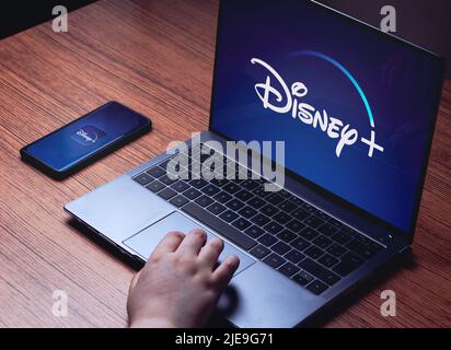 Logo Disney+ sugli schermi di notebook e smartphone. Scrivania in legno con laptop e smartphone in streaming Disney+. Foto Stock