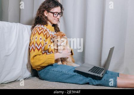 Donna in un maglione giallo e occhiali lavora a casa in un computer portatile seduto su tappeto con il suo gatto rosso, lavoro remoto da casa, freelance. Foto Stock