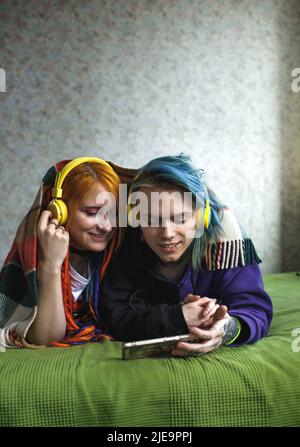 Ritratto di una giovane coppia di punk tatuati di una ragazza e di un ragazzo con capelli lunghi tinti intrecciati, giacendo su un grande letto verde. Ascoltano musica. Lar. Giallo Foto Stock