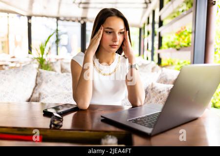 Donna stressata usando il laptop al caffè, seduto al tavolo con blocco note e tazza di caffè da solo, toccando i suoi templi, sperimentando burnout professionale, h Foto Stock