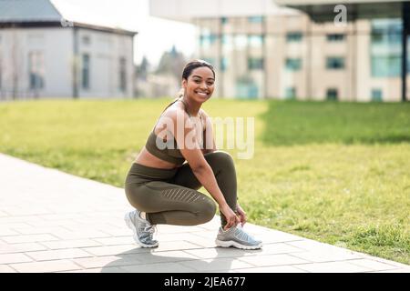 Gioiosa giovane afroamericana donna legando i lacci sulle sue sneakers prima di allenarsi, jogging al parco urbano, spazio libero Foto Stock