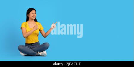 Sorridente giovane donna cinese in maglietta gialla si siede sul pavimento, punta con le dita allo spazio vuoto Foto Stock