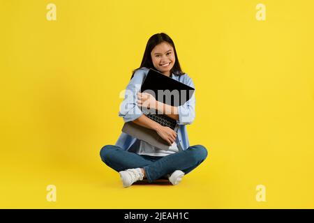 Allegro felice giovane studentessa asiatica con sindrome di Down seduta sul pavimento, abbraccio laptop Foto Stock