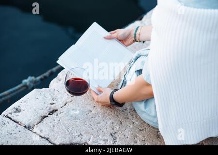 Donna in abiti blu chiaro è seduta sul molo con libro nelle mani, c'è un bicchiere di vino rosso accanto a lei. Foto Stock