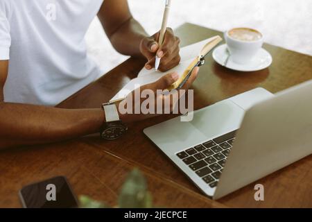 Uomo multiculturale unrecogniable che usa il laptop, prendendo le note nel libro degli esercizi, bere il caffè durante lo studio di tirocinio Foto Stock