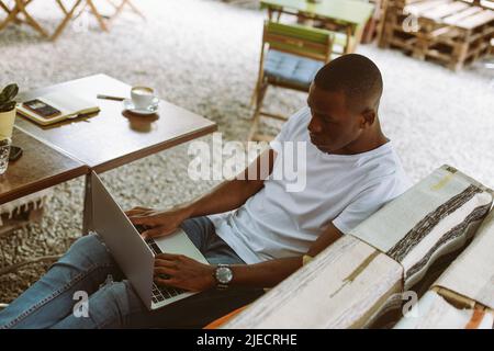 Uomo multirazziale concentrato con computer portatile seduto su panca con gambe in Street coffee shop. Preparazione al webinar online Foto Stock