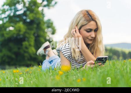 Giovane ragazza europea sdraiata sull'erba e digitando nel suo telefono. Full shot all'aperto. Foto di alta qualità Foto Stock