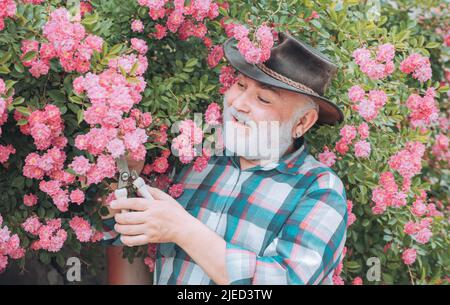 Ritratto di bell'uomo vecchio barbuto su sfondo primaverile. La vita di paese. Piantando fiori. Felice agricoltore in cappello cowboy divertirsi sul campo. Foto Stock