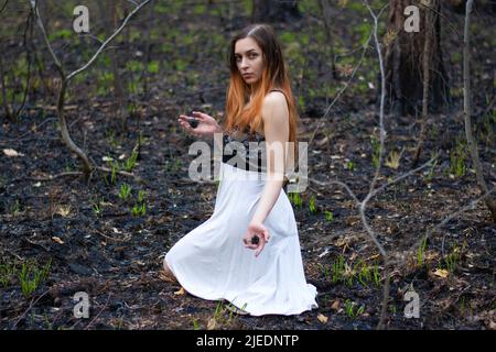 una ragazza con capelli rossi in una foresta nera bruciata. una donna tiene i coni di abete bruciato nelle sue mani. problemi di ecologia e protezione ambientale Foto Stock