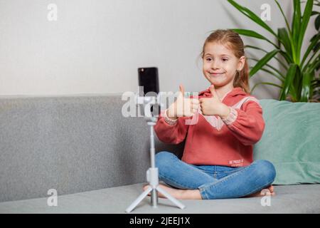 Piccola bionda blogger ragazza, una persona influente, comunica con gli abbonati, conduce una trasmissione dal vivo con un gatto, guarda lo schermo dello smartphone Foto Stock