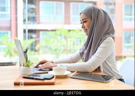Attraente giovane asiatico musulmano libera o donna d'affari a distanza che lavora nel caffè, utilizzando un computer portatile. Foto Stock