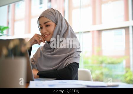 Ragazza musulmana allegra che indossa hijab siede alla sua scrivania d'ufficio, guardando lo schermo del laptop, usando il computer portatile, progettando il suo progetto di vendita. Foto Stock