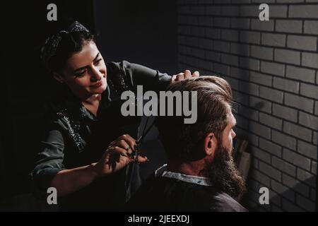 Giovane donna barbiere che fa il taglio di capelli dell'uomo barbuto in barbiere. Cura di sé, bellezza maschile. Foto Stock