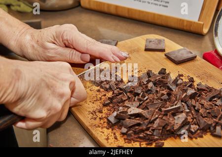 Lo chef taglia il cioccolato amaro a pezzi sul pannello di legno in cucina Foto Stock