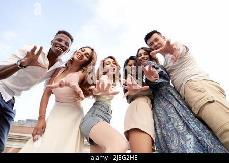 Vista ad angolo basso di un gruppo di amici multietnici che guardano la macchina fotografica e sorridono mentre si posano insieme all'aperto. Concetto di amicizia. Foto Stock