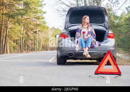 Triangolo di emergenza rosso con auto e donna seduta nel bagagliaio con thermos in background in attesa di aiuto. Macchina rotta sulla strada Foto Stock