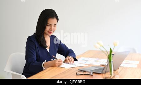 Giovane donna d'affari professionista che scrive il programma di affari sul blocco note e usando il laptop al suo posto di lavoro Foto Stock