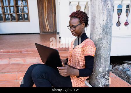 Giovane studentessa africana con occhiali e computer portatile seduti sul portico preparandosi per il suo esame Foto Stock