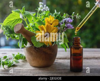 Goccia d'olio essenziale che cade in una bottiglia di vetro, primo piano. Medicina alternativa di salute. Erbe fresche in un mortaio su un tavolo di legno, sfocatura verde natura indietro Foto Stock