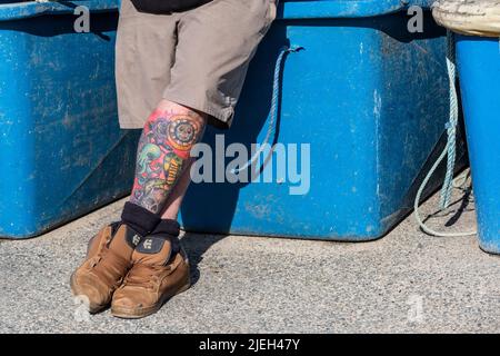 Un uomo che si rilassa contro i bidoni di stoccaggio con le gambe tatuate incrociate. Foto Stock