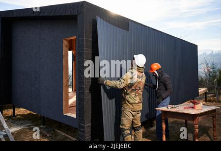 Maschio costruttori installare nero ondulato foglio di ferro usato come facciata di cottage futuro. Uomini lavoratori edificio legno telaio casa. Carpenteria e concetto di costruzione. Foto Stock