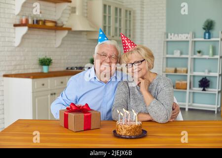 Coppia anziana in cappelli conici festosi seduti a un tavolo con un regalo e una torta con candele. Foto Stock