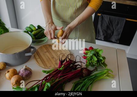 Donna croppata in grembiule pelando patate e tritare verdure fresche a bordo in cucina. Ricetta cruda per zuppa o insalata Foto Stock