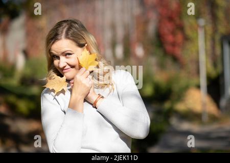 Ritratto di bionda ridente con i capelli lunghi che sbucciano giocosamente fuori da dietro il bouquet delle foglie d'autunno nelle sue mani Foto Stock