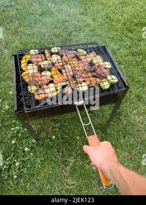 Mano di uomo tiene il traliccio sul grill con bistecche di carne e pezzi di verdure in giardino sullo sfondo del prato. Foto Stock