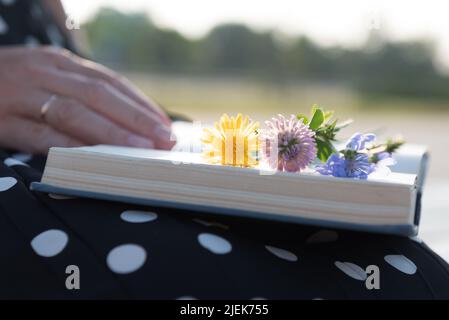 Donna sposata che legge romanzo romanticismo nel parco con fiori selvatici che giacciono sul libro, vista ritagliata. Foto Stock