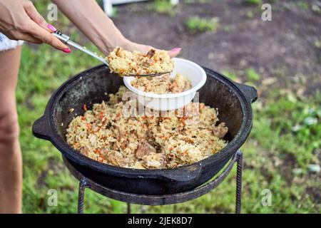 Una donna inbottona il pilaf caldo in un piatto da un calderone. Cottura del pilaf in un calderone sul fuoco nel giardino. Foto Stock