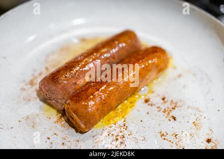 Primo piano di due salsicce finte di sostituzione della carne vegana cucinando su una padella in ceramica antiaderente in olio con buccia croccante Foto Stock