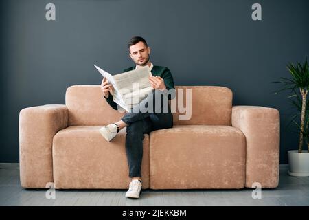 Giovane uomo sicuro che legge il giornale e le ultime notizie mentre si siede sul divano al mattino in interni moderni Foto Stock