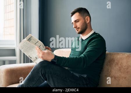 Giovane uomo sicuro che legge il giornale e le ultime notizie mentre si siede sul divano al mattino in interni moderni Foto Stock