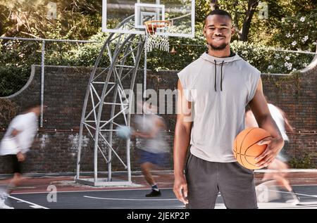 Ritratto african american uomo in piedi con una palla basket sul campo. Bel giocatore di basket maschile che tiene una palla sportiva mentre i suoi amici e. Foto Stock