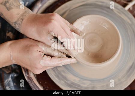 Panoramica delle mani di giovani artigiani che formano la forma di caraffa di argilla o pentola che ruota su ceramica mentre creatina terracotta Foto Stock
