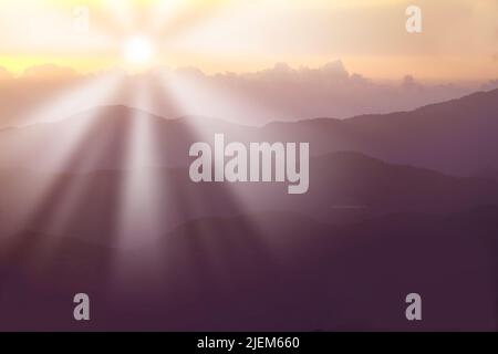 Sole che sorge dietro le montagne in estate. Raggi di sole che causano la luce sulle colline in un ambiente panoramico. Pendii forestali della catena montuosa. Disco luminoso Foto Stock