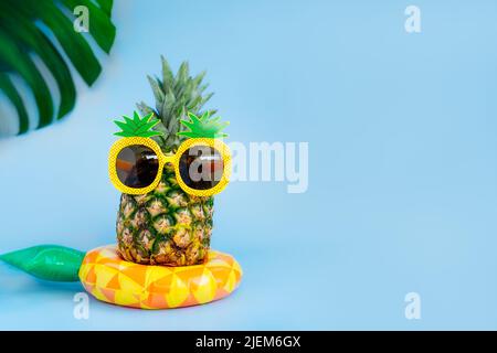 Ananas divertente in un bicchiere di ananas a forma di ananas sul giallo gonfiabile anello su sfondo blu. Estate in stile tropicale sulle vibrazioni della spiaggia. Esotico fr Foto Stock