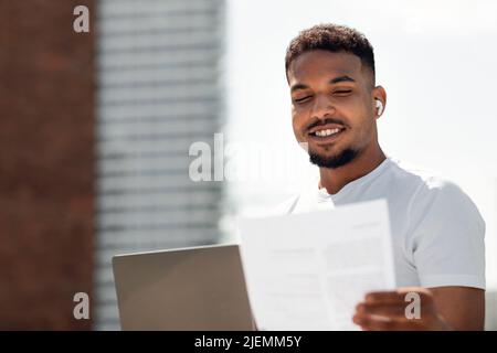 Uomo alla moda afroamericano che lavora su laptop all'aperto e controllo di documenti commerciali, spazio copia Foto Stock