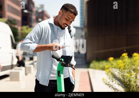 Servizi di condivisione scooter elettrici push. Giovane ragazzo nero che noleggi e-scooters in città, pagando online con smartphone Foto Stock