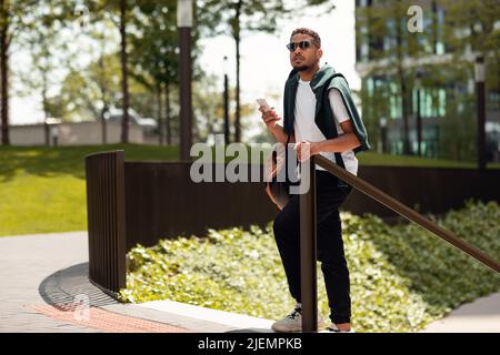 Bel ragazzo afroamericano che usa il navigatore sul cellulare o chattando in social network mentre cammina nell'area urbana Foto Stock