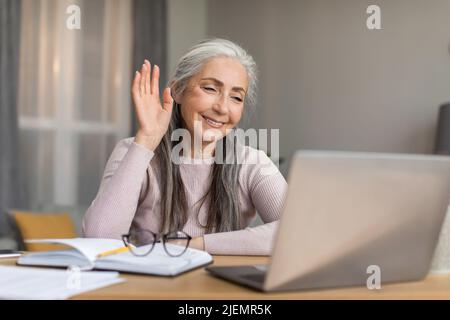 Felice sorridente donna europea anziana con capelli grigi che agita la mano al computer ha ottenuto riunione a distanza Foto Stock