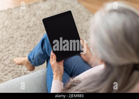 Vecchia signora europea con capelli grigi guarda il tablet con schermo vuoto, navigare in internet all'interno della camera Foto Stock