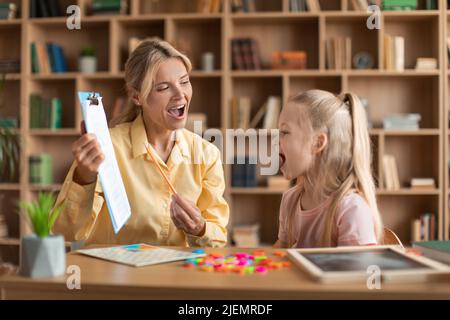 Cheerful il terapista femminile di discorso che cura i problemi e gli impedimenti del bambino, lettere imparanti della ragazza piccola durante la lezione Foto Stock