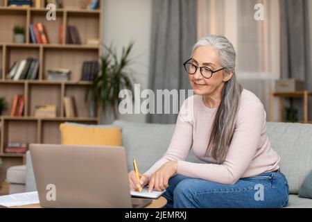 Concentrato europeo insegnante mature signora con capelli grigi in occhiali lavora con il computer, hanno riunione a distanza Foto Stock