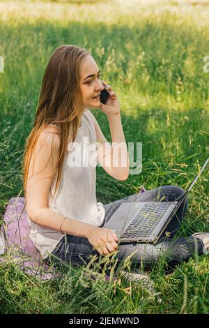 la ragazza si siede sull'erba e lavora a un portatile. parlando al telefono. freelance. selfeducation. il concetto di apprendimento remoto e di lavoro all'aperto Foto Stock