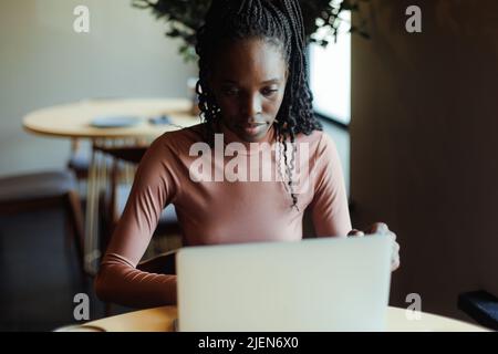 Ritratto di giovane donna afro-americana concentrata seduta al tavolo di legno beige in caffetteria, lavorando a scrivere su un computer portatile. Foto Stock