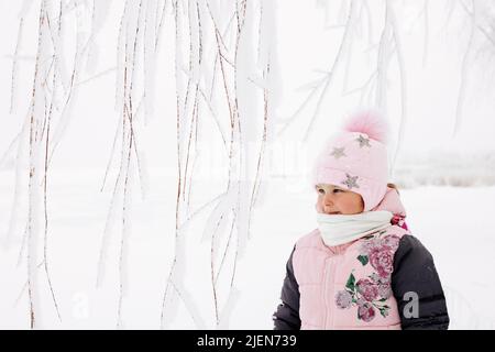 Rametti di alberi ricoperti di neve con ragazza ghiacciata con guance rosse che guardano via vestito con caldi vestiti invernali con campo nevoso sullo sfondo. Passeggiata invernale Foto Stock