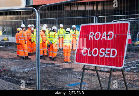 Red Road ha chiuso il cartello con i lavoratori delle costruzioni riuniti per l'istruzione da parte del responsabile del sito. Giacche e abiti ad alta visibilità, cappelli rigidi e stivali. Foto Stock