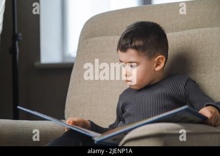 Ragazzino che legge un libro mentre si siede sul divano, a casa. Istruzione e sviluppo precoce dei bambini. Foto Stock